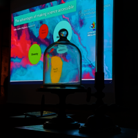una cupola di vetro su un supporto di fronte a uno schermo di proiezione. La cupola è trasparente e permette di vedere lo schermo di proiezione dall'interno. Lo schermo mostra una presentazione con il titolo "I vantaggi di rendere la scienza accessibile"